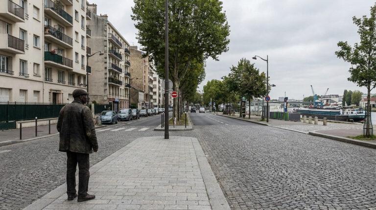 Un homme debout sur un trottoir pavé le long d'une rue bordée d'immeubles résidentiels et de la Seine à Ivry.